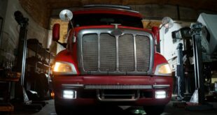 Front view of a red truck in a dimly lit automotive repair shop with a focus on the grill and headlights.