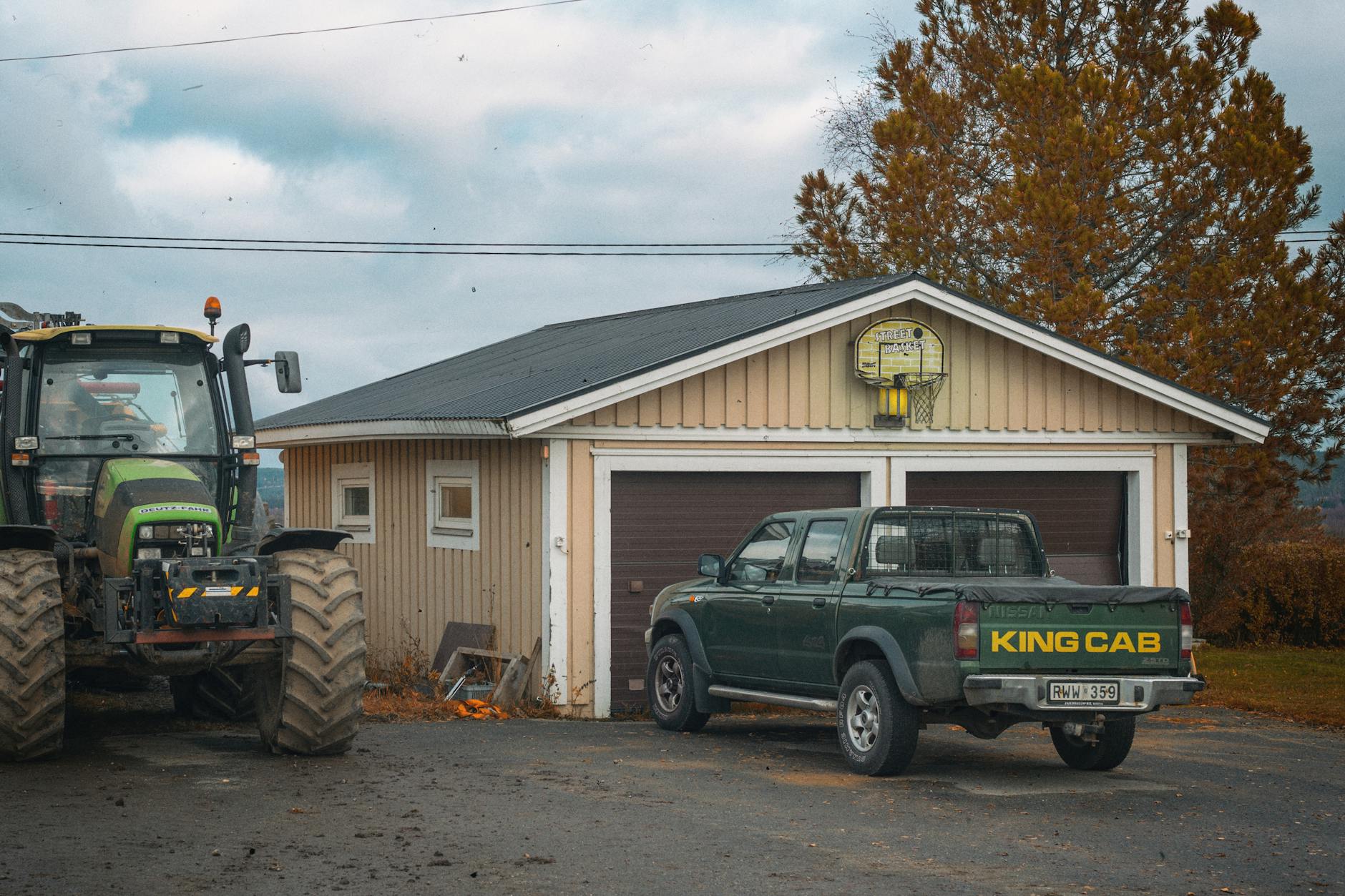 A rural scene in Hedenäset, Sweden featuring a tractor and King Cab pickup truck parked by a garage.