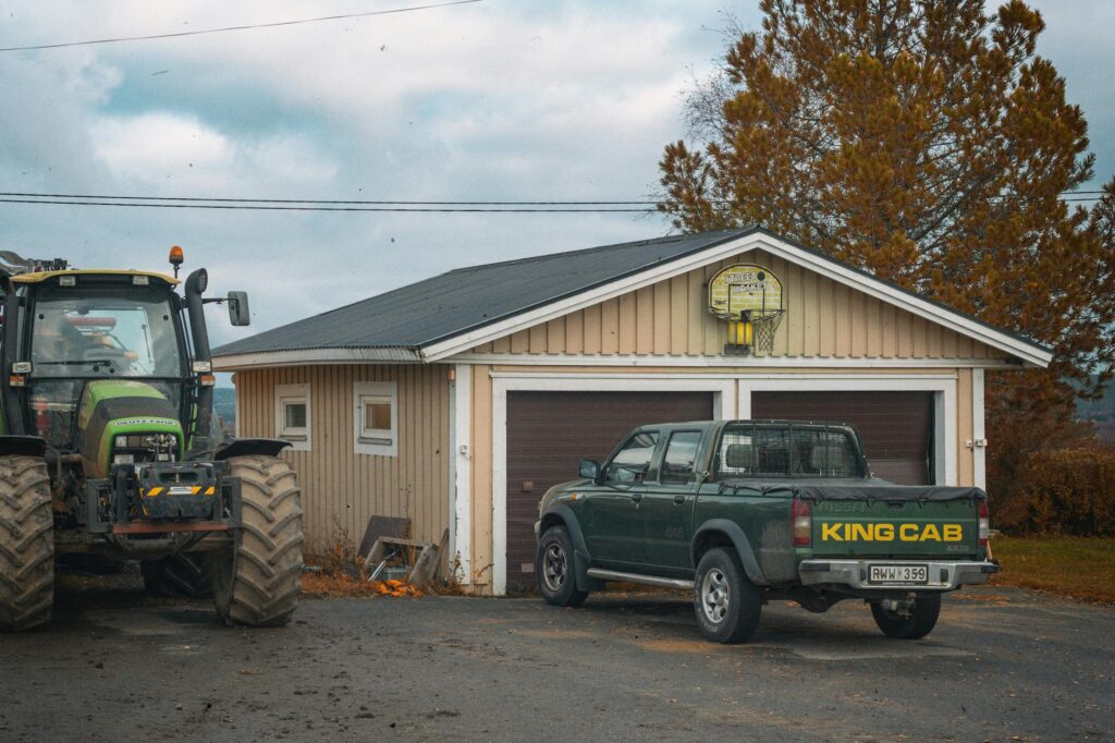 A rural scene in Hedenäset, Sweden featuring a tractor and King Cab pickup truck parked by a garage.
