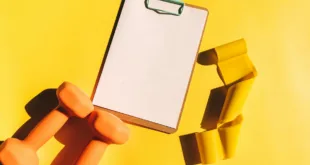 Bright flat lay of orange dumbbells, clipboard, and yellow resistance band on yellow background.