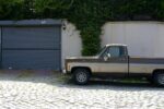 A vintage brown pickup truck parked in front of a suburban garage with greenery.