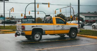 A yellow pickup truck parked in a lot with a hiring sign on a rainy day.