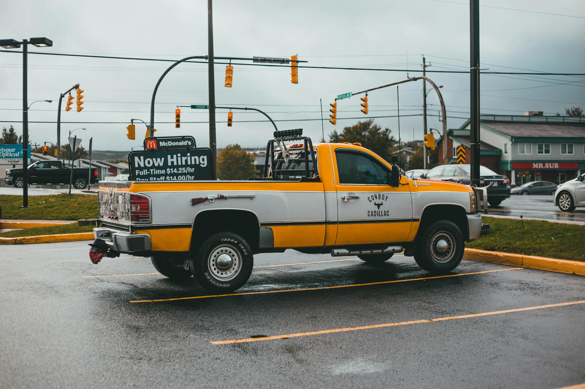 A yellow pickup truck parked in a lot with a hiring sign on a rainy day.