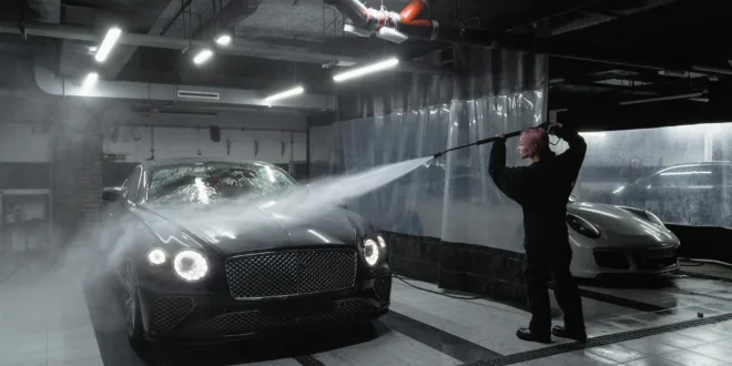 A person washing a black luxury car in a dimly lit garage using a pressure washer.