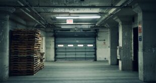Empty industrial warehouse featuring stacked pallets and a closed garage door under fluorescent lighting.