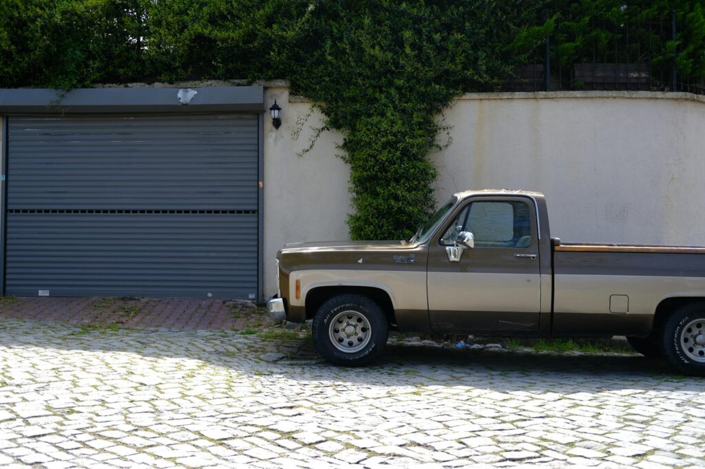 A vintage brown pickup truck parked in front of a suburban garage with greenery.