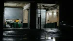Dark industrial garage at night with open doors showing bins and equipment.
