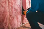 Crop unrecognizable worker in gloves sitting on haunches and insulating with pink stone wool