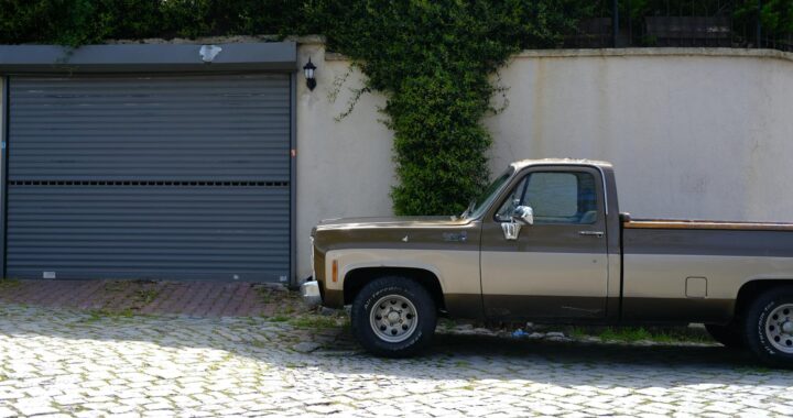 A vintage brown pickup truck parked in front of a suburban garage with greenery.