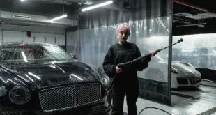 A woman with pink hair uses a pressure washer on a luxury car in an indoor garage setting.